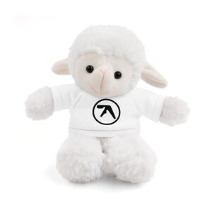 May include: A white plush lamb toy wearing a white t-shirt with a black circular logo. The lamb has a fluffy, textured body, small ears, and black eyes. The toy is designed to sit upright.