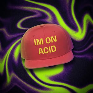 May include: A red baseball cap with a flat brim and a structured crown. The front of the cap features the text IM ON ACID printed in bold yellow capital letters. The design is centered on the front panels of the hat, which has a solid red color.