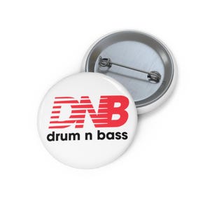 Może przedstawiać: Biała przypinka z czerwonym napisem "DNB" i czarnymi słowami "drum n bass". Przypinka ma metalową szpilkę z tyłu. Przypinka jest okrągła i ma błyszczące, metaliczne wykończenie.