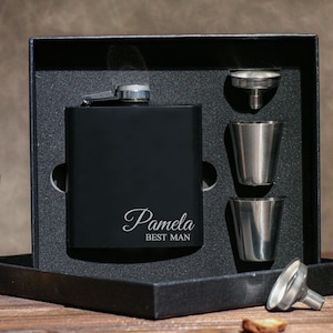 Puede incluir: Un juego de regalo de petaca negra con el nombre "Pamela" y "BEST MAN" en escritura blanca. El juego incluye dos vasos de chupito y dos embudos. La petaca es rectangular y tiene una tapa plateada.