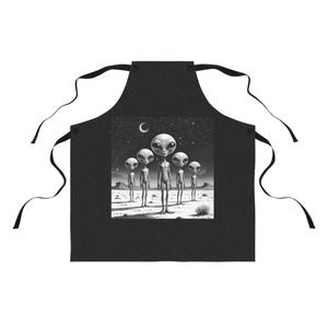Puede incluir: Delantal negro con una ilustración en escala de grises de cinco extraterrestres en un paisaje desértico bajo un cielo estrellado y una luna creciente. El delantal tiene lazos negros.
