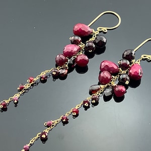 Op de afbeelding: Een paar lange, hangende oorbellen met een cluster van rode en donkerrode edelstenen. De oorbellen zijn gemaakt van goud en hebben een delicaat kettingontwerp.
