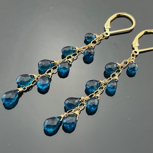 May include: A pair of gold-toned dangle earrings featuring multiple teardrop-shaped, faceted, blue gemstones. The gemstones are suspended from delicate gold chains, creating a cascading effect. The earrings have a lever-back closure.