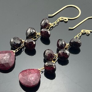 Op de afbeelding: Een paar gouden oorbellen met een hangende traanvormige robijn en meerdere kleinere, ronde, donkerrode edelstenen.