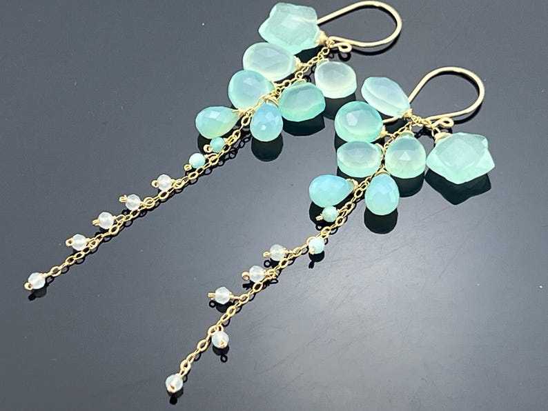 May include: Elegant gold-tone earrings featuring clusters of light blue and clear gemstones. The earrings have a cascading design with various shapes of gemstones, including teardrops and faceted stones, suspended from delicate chains.