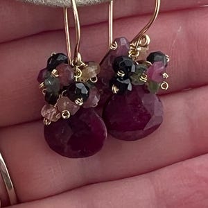 May include: Gold-tone earrings featuring clusters of small, colorful gemstones. The earrings have a teardrop-shaped, faceted, deep red ruby at the bottom. The gemstones include black, pink, and green stones.