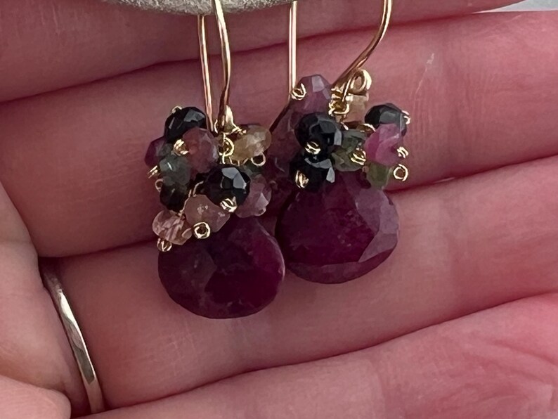 May include: Gold-tone earrings featuring clusters of small, colorful gemstones. The earrings have a teardrop-shaped, faceted, deep red ruby at the bottom. The gemstones include black, pink, and green stones.