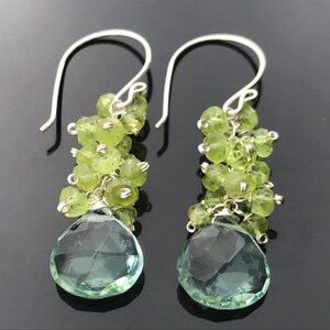 Op de afbeelding: Zilveren oorhangers met een traanvormige lichtgroene edelsteen en een cluster van kleinere lichtgroene edelstenen.