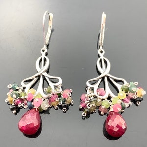 Op de afbeelding: Zilveren oorhangers met een cluster van kleurrijke edelstenen, waaronder roze, groen en blauw, en een druppelvormige robijn hanger.