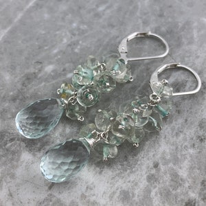May include: A pair of silver dangle earrings featuring light blue aquamarine gemstones. The earrings have a cluster of faceted aquamarine stones and a teardrop-shaped aquamarine pendant.