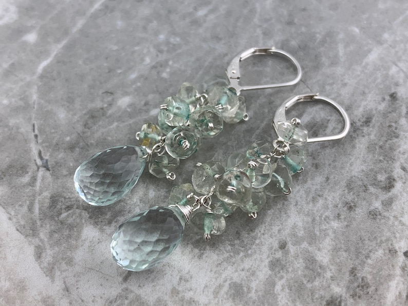 May include: A pair of silver dangle earrings featuring light blue aquamarine gemstones. The earrings have a cluster of faceted aquamarine stones and a teardrop-shaped aquamarine pendant.