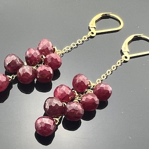 May include: Gold-coloured earrings featuring clusters of faceted, deep red gemstones. The earrings have a lever-back clasp and hang from a fine chain. The gemstones are arranged in a cascading design, creating a stylish accessory.