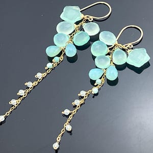 May include: A pair of gold-toned dangle earrings featuring clusters of light blue and aqua-colored gemstones. The earrings have a cascading design with small white beads strung on delicate chains.