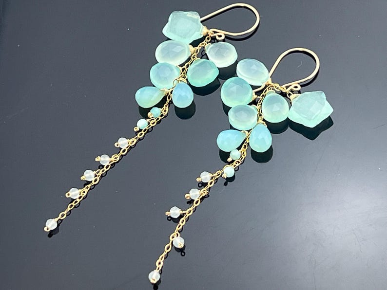 May include: A pair of gold-toned dangle earrings featuring clusters of light blue and aqua-colored gemstones. The earrings have a cascading design with small white beads strung on delicate chains.