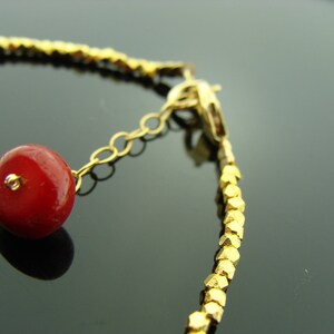 Op de afbeelding: Een gouden armband met een rode kralenbedel. De armband is gemaakt van kleine, gouden, gefacetteerde kralen. De bedel is met een gouden ketting aan de armband bevestigd.