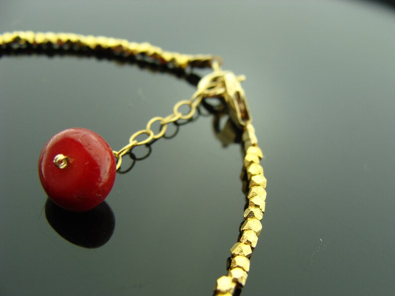 Op de afbeelding: Een gouden armband met een rode kralenbedel. De armband is gemaakt van kleine, gouden, gefacetteerde kralen. De bedel is met een gouden ketting aan de armband bevestigd.