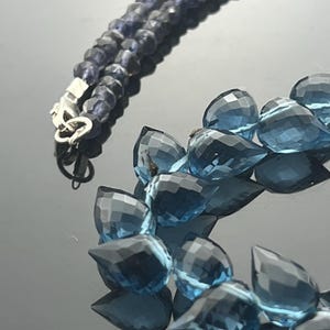 May include: A close-up of a necklace with two strands of blue gemstones. One strand features small, round, dark blue beads, while the other has larger, teardrop-shaped, light blue gems. A silver clasp connects the strands.