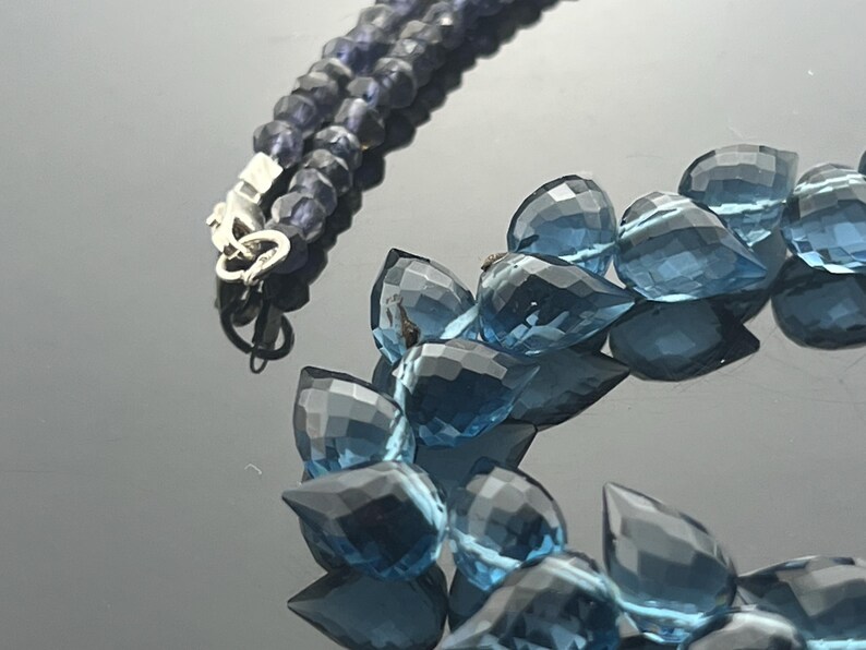 May include: A close-up of a necklace with two strands of blue gemstones. One strand features small, round, dark blue beads, while the other has larger, teardrop-shaped, light blue gems. A silver clasp connects the strands.