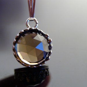 May include: A round pendant with a faceted, smoky-colored gemstone. The gem is set in a silver-toned, ornate bezel. The pendant hangs from a silver bail. The background is a gradient of gray and black.