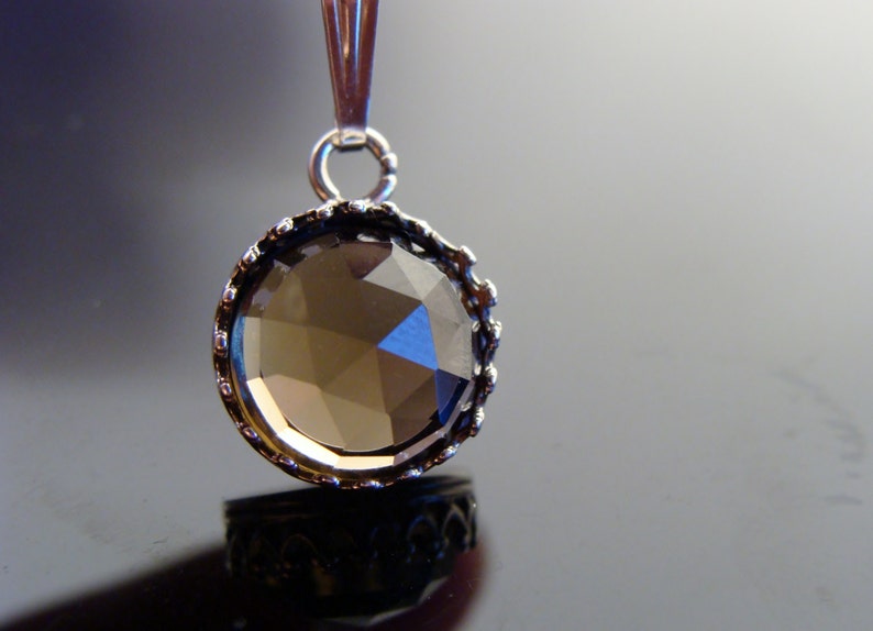 May include: A round pendant with a faceted, smoky-colored gemstone. The gem is set in a silver-toned, ornate bezel. The pendant hangs from a silver bail. The background is a gradient of gray and black.