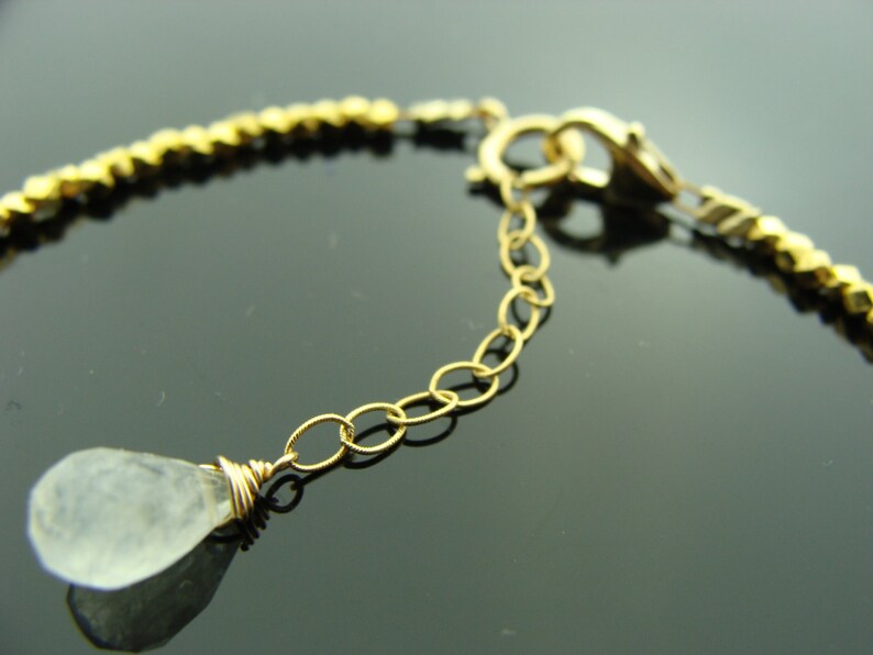 May include: A gold chain bracelet with a clear teardrop-shaped gemstone pendant. The bracelet has a lobster clasp closure.