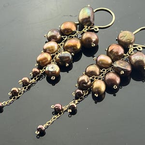 May include: A pair of gold dangle earrings with brown freshwater pearls. The pearls are arranged in a cascading cluster, creating a delicate and elegant look.