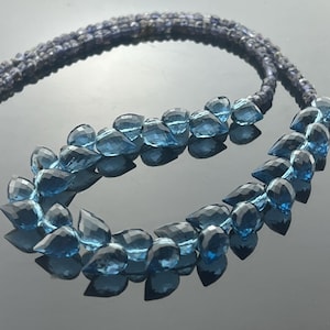 May include: A necklace featuring two strands of small, dark blue beads, with a series of larger, faceted, teardrop-shaped blue gemstones. The necklace has a silver clasp and is displayed on a reflective surface.