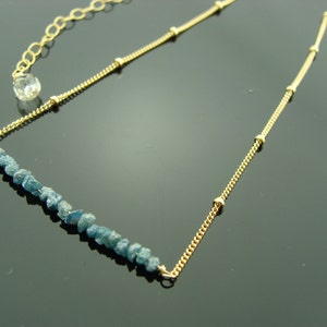 May include: A gold chain necklace with a row of blue rough diamonds. The chain has small gold beads.