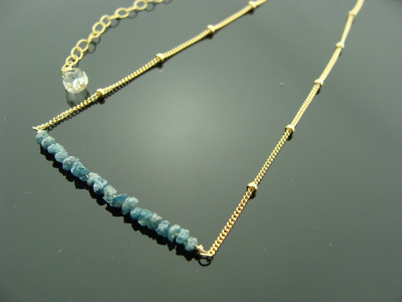 May include: A gold chain necklace with a row of blue rough diamonds. The chain has small gold beads.