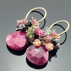 May include: A pair of gold-colored earrings with a cluster of colorful gemstones. Each earring features a large, faceted, teardrop-shaped pink stone and smaller, round gemstones in various colors.