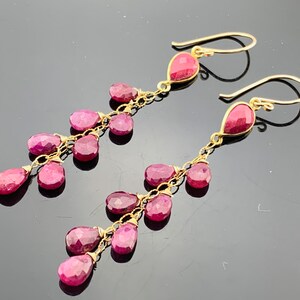 May include: Gold dangle earrings featuring multiple pink ruby gemstones. The earrings are teardrop shaped and connected by a delicate chain.