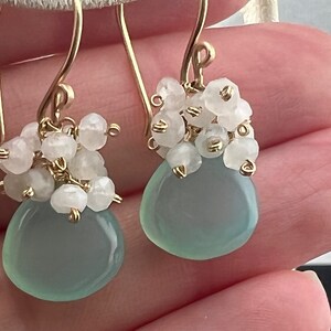 May include: A pair of gold earrings featuring a light blue gemstone and white beads. The earrings are dangle style and have a delicate design.