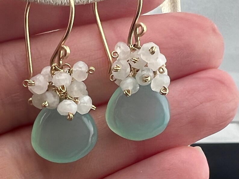 May include: A pair of gold earrings featuring a light blue gemstone and white beads. The earrings are dangle style and have a delicate design.