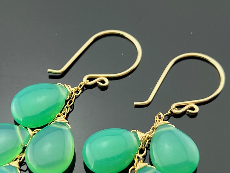 May include: A pair of gold dangle earrings with green gemstone drops. The earrings feature a delicate chain and multiple teardrop-shaped stones.