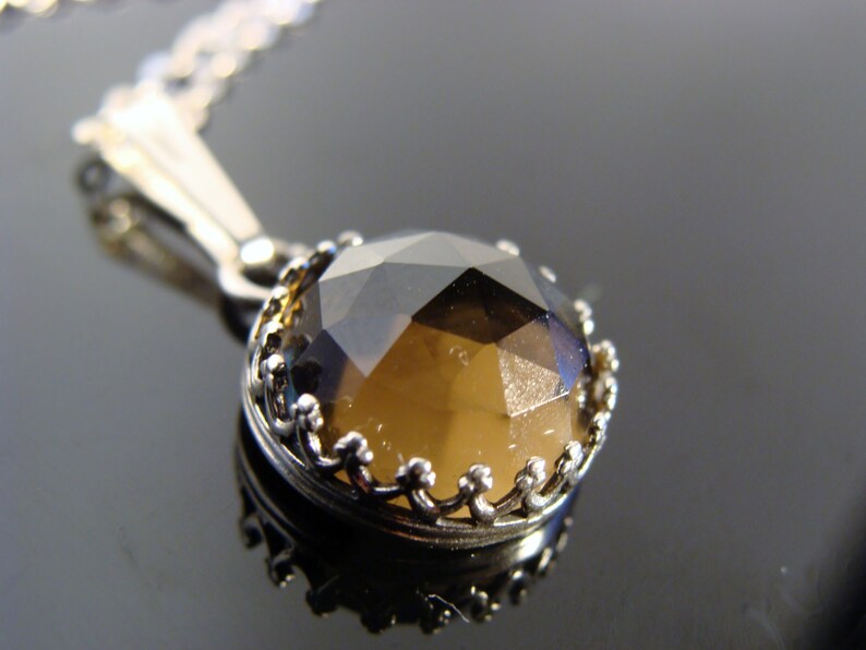 May include: A silver pendant necklace featuring a faceted, round, smoky quartz gemstone. The gemstone is set in a decorative, crown-like bezel. The chain is delicate and silver, creating an elegant and classic jewelry piece.