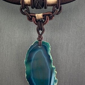 Pu&ograve; includere: Una collana di corda marrone scuro con un ciondolo in agata verde. Il ciondolo &egrave; una fetta di agata con un motivo bianco e verde.