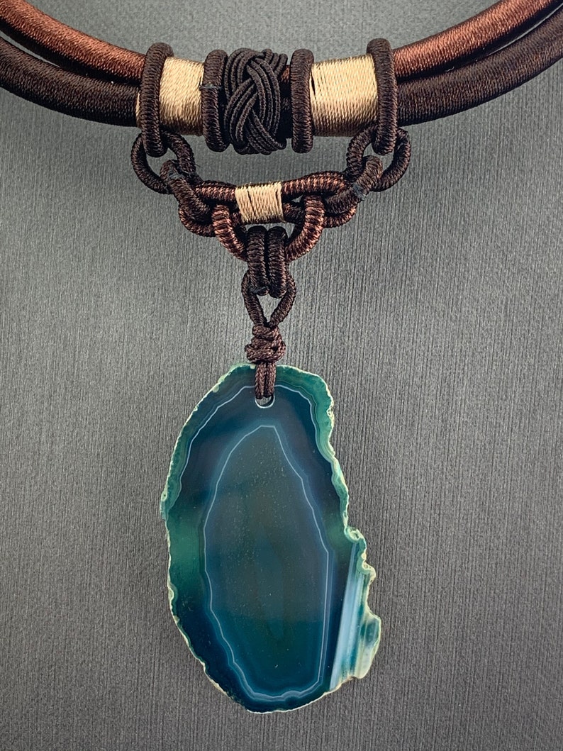 Pu&ograve; includere: Una collana di corda marrone scuro con un ciondolo in agata verde. Il ciondolo &egrave; una fetta di agata con un motivo bianco e verde.