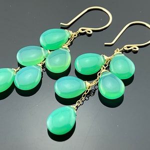 May include: A pair of gold dangle earrings with multiple teardrop-shaped green gemstones. The earrings are connected by a delicate gold chain.