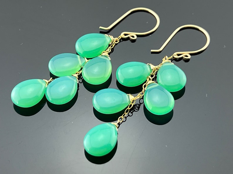 May include: A pair of gold dangle earrings with multiple teardrop-shaped green gemstones. The earrings are connected by a delicate gold chain.