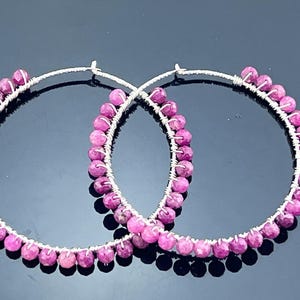 May include: A pair of large hoop earrings crafted from thin silver-toned wire. Each hoop is adorned with a continuous row of small, faceted, round, deep pink beads that are individually wrapped and secured along the entire circumference of the wire.
