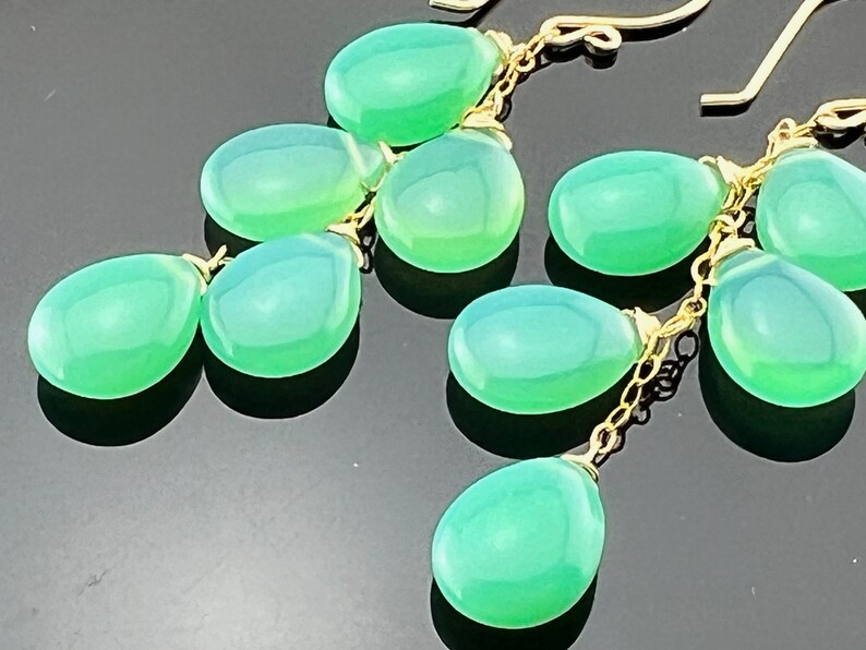 May include: A pair of green gemstone dangle earrings with gold chain links. The earrings feature multiple teardrop-shaped gemstones.