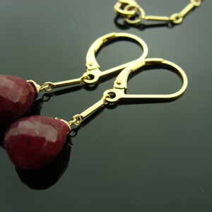 May include: Gold earrings with red gemstone drops. The earrings have a simple design with a small gold hoop and a dangling gemstone.