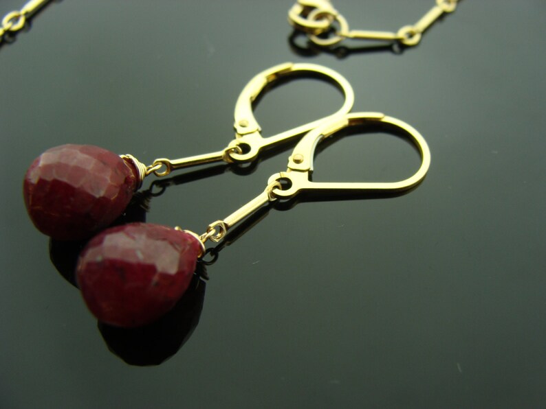May include: Gold earrings with red gemstone drops. The earrings have a simple design with a small gold hoop and a dangling gemstone.