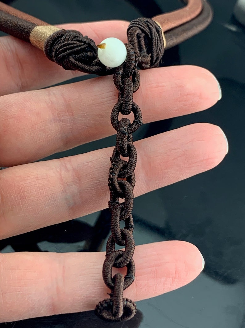 Pu&ograve; includere: Una collana di corda marrone con un ciondolo bianco e un'estensione a catena marrone.