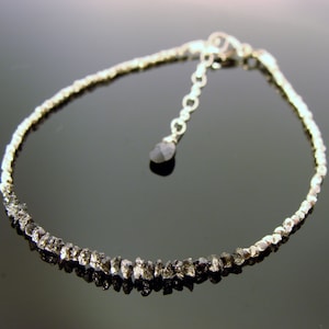 May include: A silver chain bracelet with a small black diamond pendant. The bracelet is made of small, rough black diamonds.
