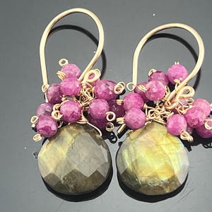 May include: A pair of gold-tone wire earrings featuring faceted, teardrop-shaped labradorite stones suspended below clusters of small, round, deep pink ruby beads. The earrings are attached to simple gold-tone ear wires.