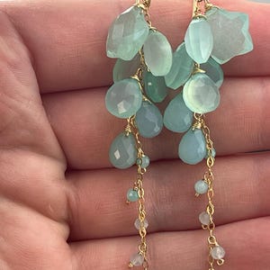 May include: Elegant dangle earrings featuring clusters of light blue and green gemstone beads. The earrings have a gold-colored chain and wire accents, with various shapes and sizes of beads.