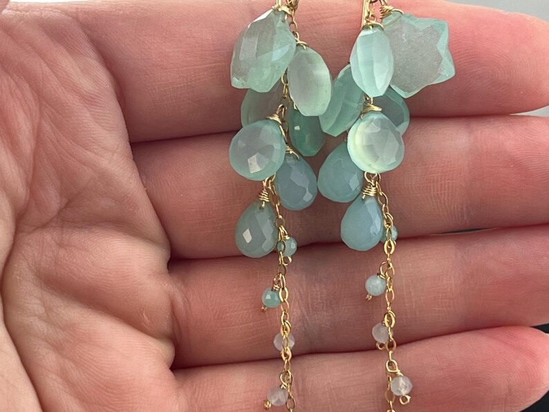 May include: Elegant dangle earrings featuring clusters of light blue and green gemstone beads. The earrings have a gold-colored chain and wire accents, with various shapes and sizes of beads.