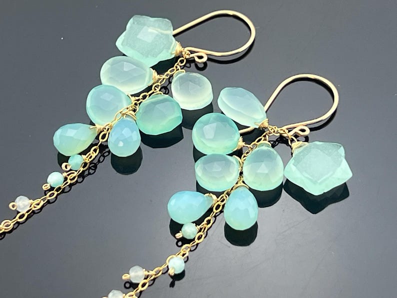 May include: A pair of gold-tone earrings featuring clusters of light blue and green gemstones. The earrings have a delicate chain design with various shapes of gemstones, including teardrops and irregular shapes.