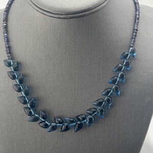 May include: A necklace featuring a leaf-like design. The necklace has dark blue, faceted gemstone leaves strung on a light blue cord. The necklace is displayed on a gray mannequin bust.